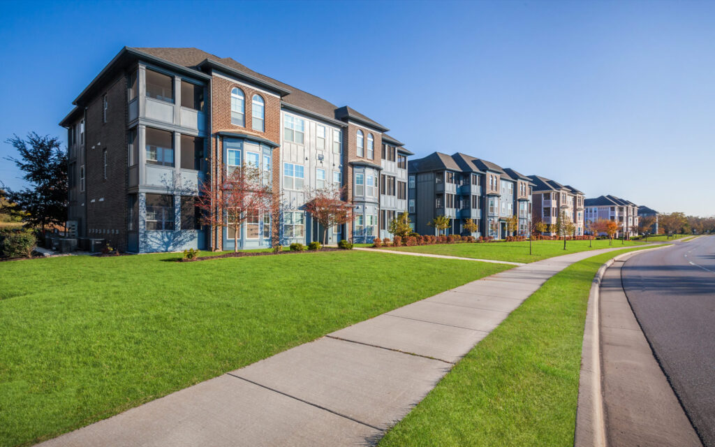 A row of modern, three-story townhouses with large windows and balconies lines a clean sidewalk beside a wide street under a clear blue sky, with neatly trimmed grass and small trees in front.