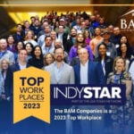 A large group of professionally dressed people poses together for a photo. In front, banners read Top Workplaces 2023, INDYSTAR, and The BAM Companies is a 2023 Top Workplace.
