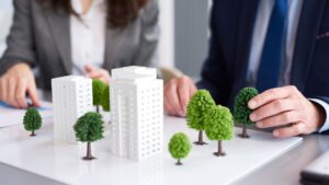 Two people in business attire discuss a city planning model, featuring miniature white buildings and green trees arranged on a table. The focus is on the model while their faces are out of focus.