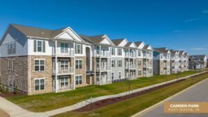 A row of modern, three-story apartment buildings with balconies and stone accents, set along a tidy street under a clear blue sky. A sign at the bottom right reads Camden Park, Fort Wayne, IN.