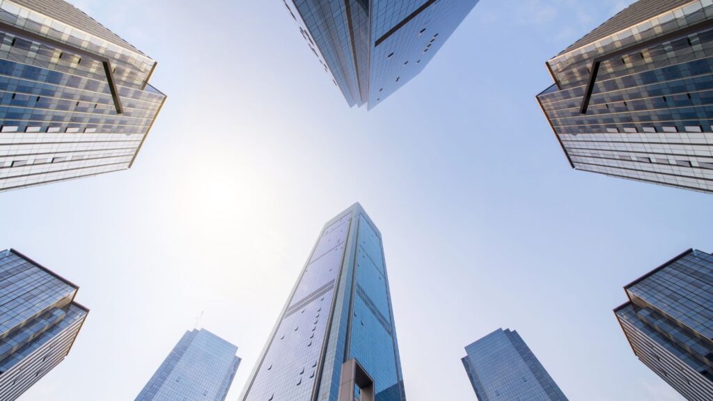 Tall skyscrapers with glass facades surround the view, seen from the ground looking up toward a clear blue sky and bright sunlight in the background.