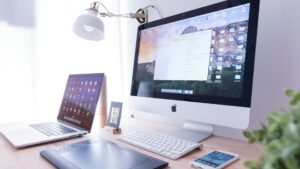 A tidy workspace with a desktop iMac, open MacBook, wireless keyboard and mouse, graphics tablet, desk lamp, smartphone, and a small potted plant on a light wooden desk.
