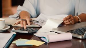 A person uses a calculator while holding a pen and reviewing documents at a desk, with a laptop, coffee cup, notepad, and colorful sticky notes scattered nearby.