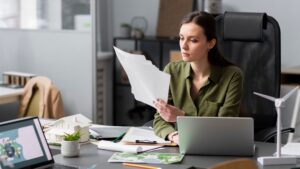 A woman sits at a desk in an office, looking at papers. She is surrounded by a laptop, documents, architectural models, and a small wind turbine model, suggesting she works in a field related to sustainability or design.