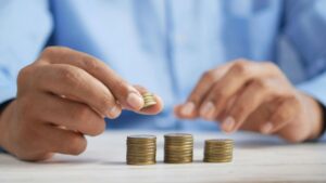 A person in a blue shirt stacks coins into three neat piles on a white table, with one hand holding a small stack and the other hand adding coins to a pile.