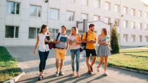 Five young adults walk together on a sunny day outside a modern building, carrying books and backpacks, and chatting cheerfully as they walk along a paved path.
