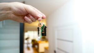 A hand holds a keyring with several keys and a black house-shaped keychain in a bright, modern home interior. The background shows a staircase and a blurred living area.