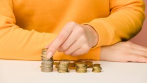 A person in a yellow sweater is stacking coins on a white surface, organizing several small piles of coins with their hand.