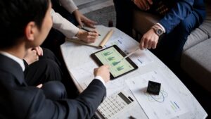 Three people in business attire sit around a table with charts, a calculator, and an iPad displaying bar graphs, engaged in a discussion and pointing at documents and the tablet.