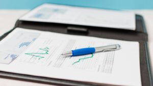 A blue and silver pen rests on printed financial documents with charts and graphs, placed on an open black folder. Another document with charts is visible in the background.