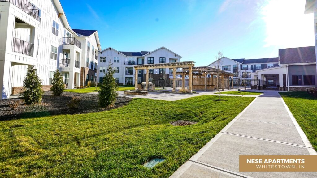 A modern apartment complex with white buildings, balconies, landscaped green lawns, a wooden pergola, and a wide sidewalk under a clear blue sky. Text reads Nese Apartments, Whitestown, IN.