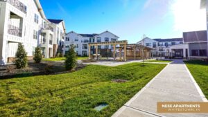 A modern apartment complex with white buildings, balconies, landscaped green lawns, a wooden pergola, and a wide sidewalk under a clear blue sky. Text reads Nese Apartments, Whitestown, IN.