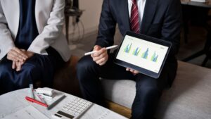 Two people in business attire sit on a couch; one holds a tablet displaying colorful bar graphs and gestures at the screen with a stylus. Office essentials like papers, a calculator, and pens are on a table nearby.