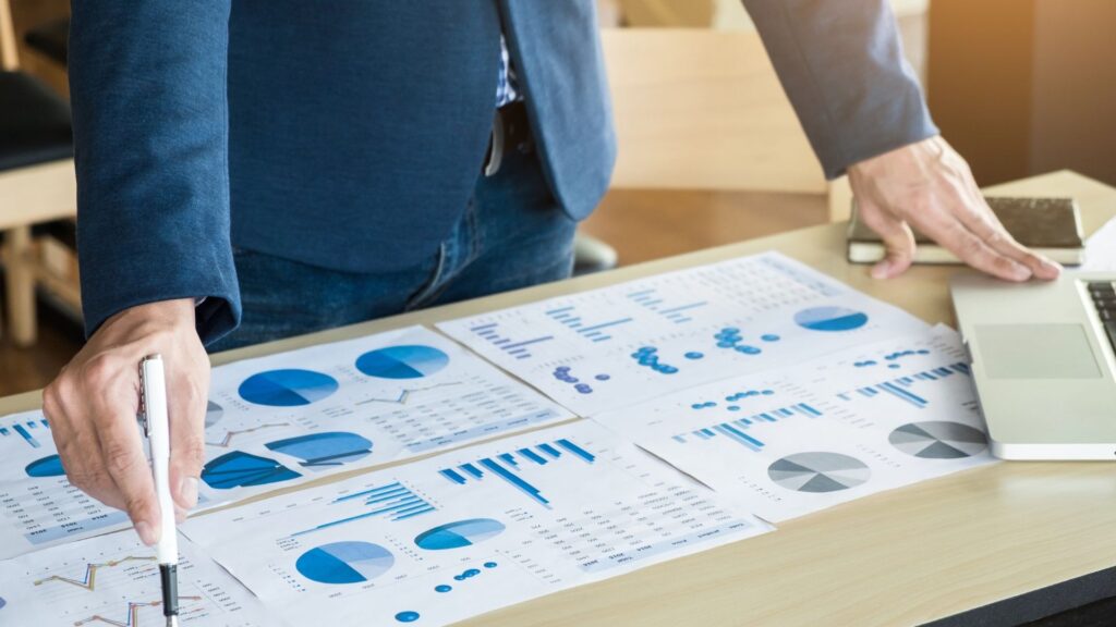 A person in a suit stands at a desk covered with printed charts and graphs, pointing at one document with a pen. A laptop and smartphone are also on the desk.