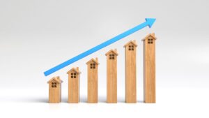 Wooden house models arranged in ascending height, forming a bar graph, with a large blue upward arrow above them, symbolizing growth or rising real estate values.
