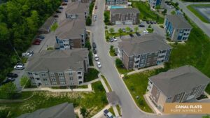Aerial view of a modern apartment complex with multiple buildings, parking lots, and greenery. Cars are parked along the streets and in parking areas. Text in the corner reads: Canal Flats, Fort Wayne, IN.