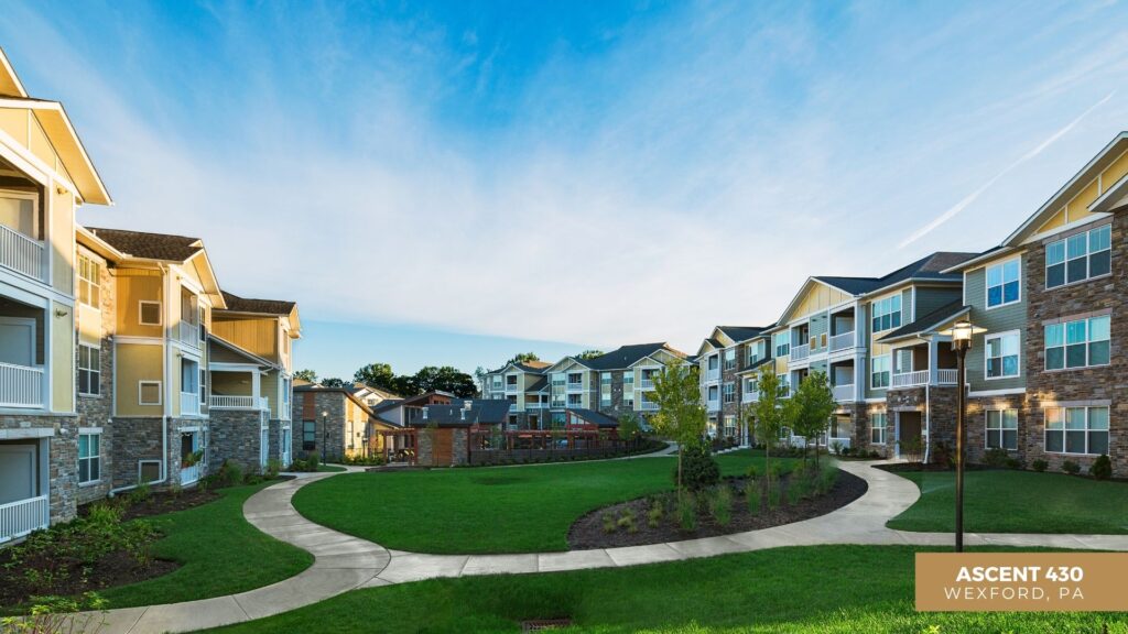 Modern apartment buildings surround a well-maintained green courtyard with winding sidewalks, landscaping, and a clear blue sky. A sign in the corner reads Ascent 430, Wexford, PA.