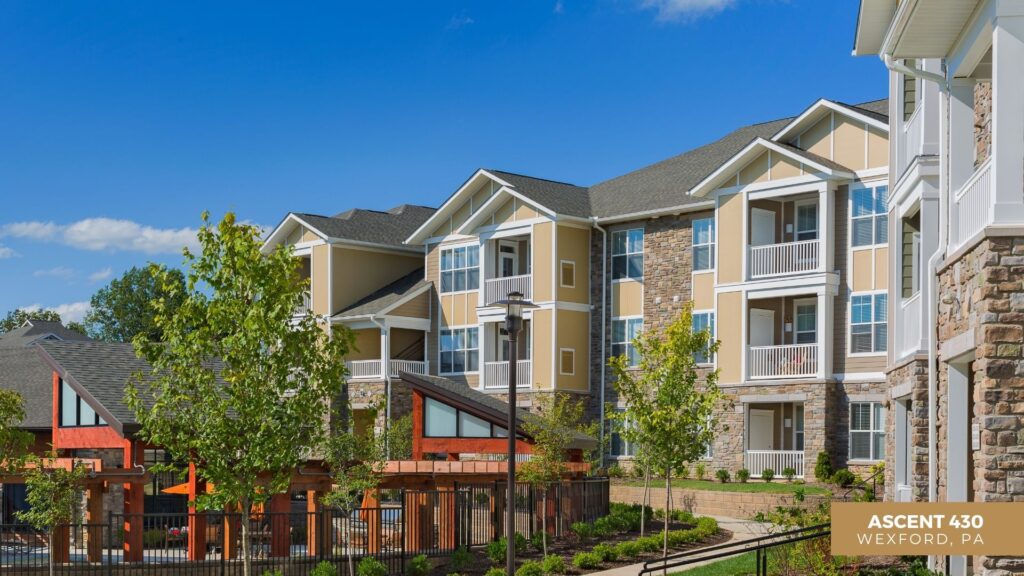 Modern apartment complex with beige siding and stone accents, featuring balconies and large windows, set against a clear blue sky. Lush landscaping and wooden pergolas are visible in the foreground. Text: Ascent 430, Wexford, PA.