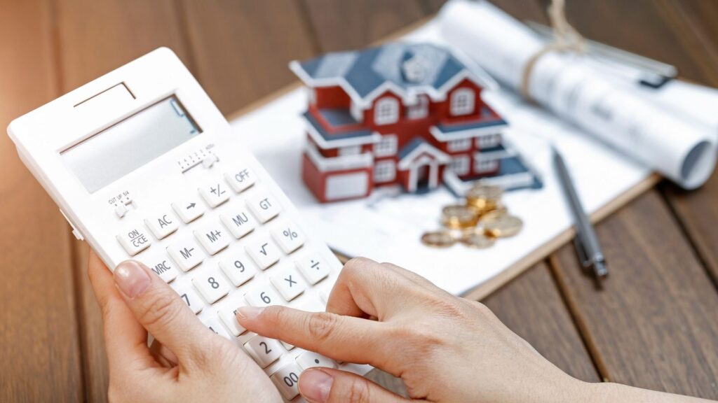 A person uses a white calculator on a wooden table with a model house, coins, rolled-up documents, and a pen in the background, symbolizing real estate or home financial planning.