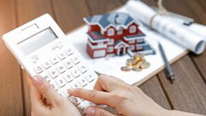 A person uses a white calculator on a wooden table with a model house, coins, rolled-up documents, and a pen in the background, symbolizing real estate or home financial planning.