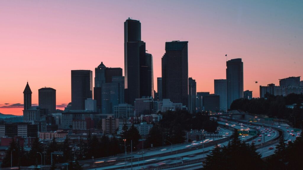 A city skyline with tall buildings silhouetted against a pink and purple sunset sky, with highways and light trails from cars in the foreground.