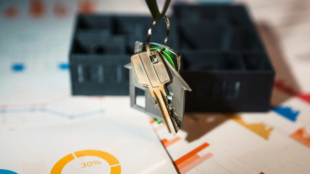 A set of house keys with a house-shaped keychain rests on documents with colorful charts and graphs, next to a small model of a house, symbolizing real estate and investment.