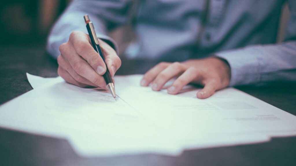 A person in a blue shirt writes on a stack of white papers with a black pen while resting their other hand on the desk.
