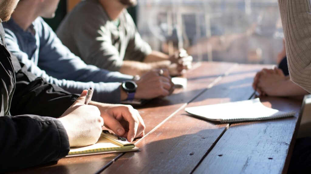 Four people sit around a wooden table, writing in notebooks and having a discussion. Sunlight shines through a nearby window, creating a bright and collaborative atmosphere. Faces are not clearly visible.