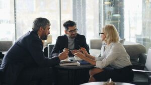 Three people in business attire sit around a small round table in a modern office, engaged in a discussion. Two men and one woman are holding pens and papers, appearing focused and collaborative.
