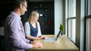 Two people stand at a wooden desk by a window, looking at a laptop. One person is typing while the other observes. There is a small potted plant on the desk and shelves in the background.