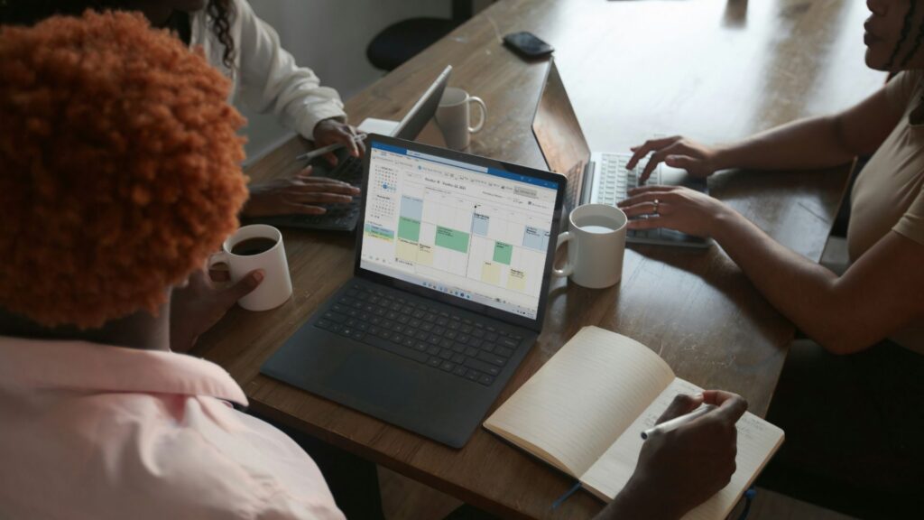 Three people sit at a wooden table with laptops, notebooks, and coffee mugs. One laptop screen displays a calendar. One person writes in a notebook while others type on keyboards, suggesting a collaborative work meeting.