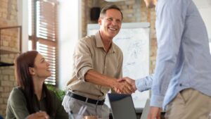 Three people in an office setting; one man is smiling and shaking hands with another person, while a woman sits nearby, watching and smiling. A whiteboard with drawings is visible in the background.
