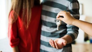 A person hands over a key to a couple standing together, with one person in a red top and the other in a striped shirt, suggesting they are receiving keys to a new home.
