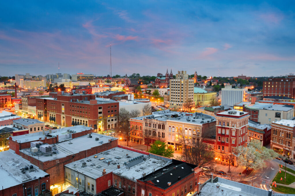 Aerial view of a cityscape at dusk, with historic brick buildings, lit streetlights, and a colorful sky transitioning from blue to pink. Trees and city landmarks are visible throughout the urban area.