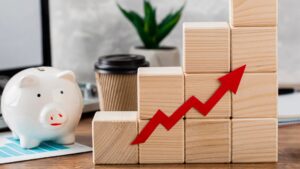 A white piggy bank, paper chart, and stacked wooden blocks with a red upward arrow symbolize financial growth. A takeaway coffee cup and a green plant are in the background on a desk.
