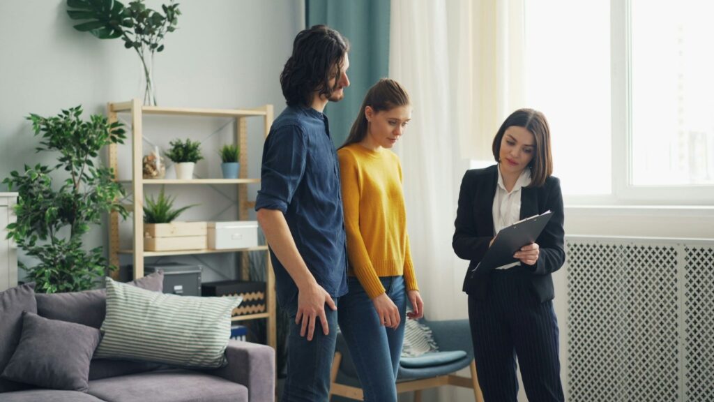 A woman in business attire shows a clipboard to a man and woman in casual clothes inside a modern living room with plants and shelves in the background.
