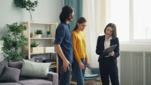 A woman in business attire shows a clipboard to a man and woman in casual clothes inside a modern living room with plants and shelves in the background.