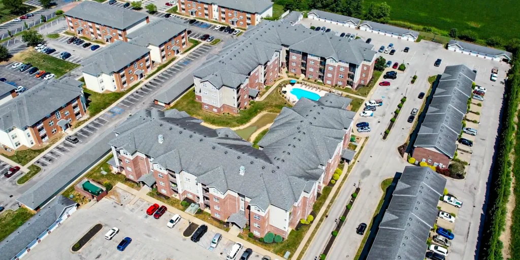 Aerial view of a large apartment complex with multiple brick buildings, parking lots with cars, a small central courtyard, and a swimming pool, surrounded by streets and greenery.