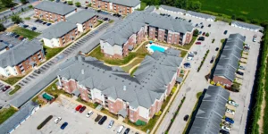 Aerial view of a large apartment complex with multiple brick buildings, parking lots with cars, a small central courtyard, and a swimming pool, surrounded by streets and greenery.