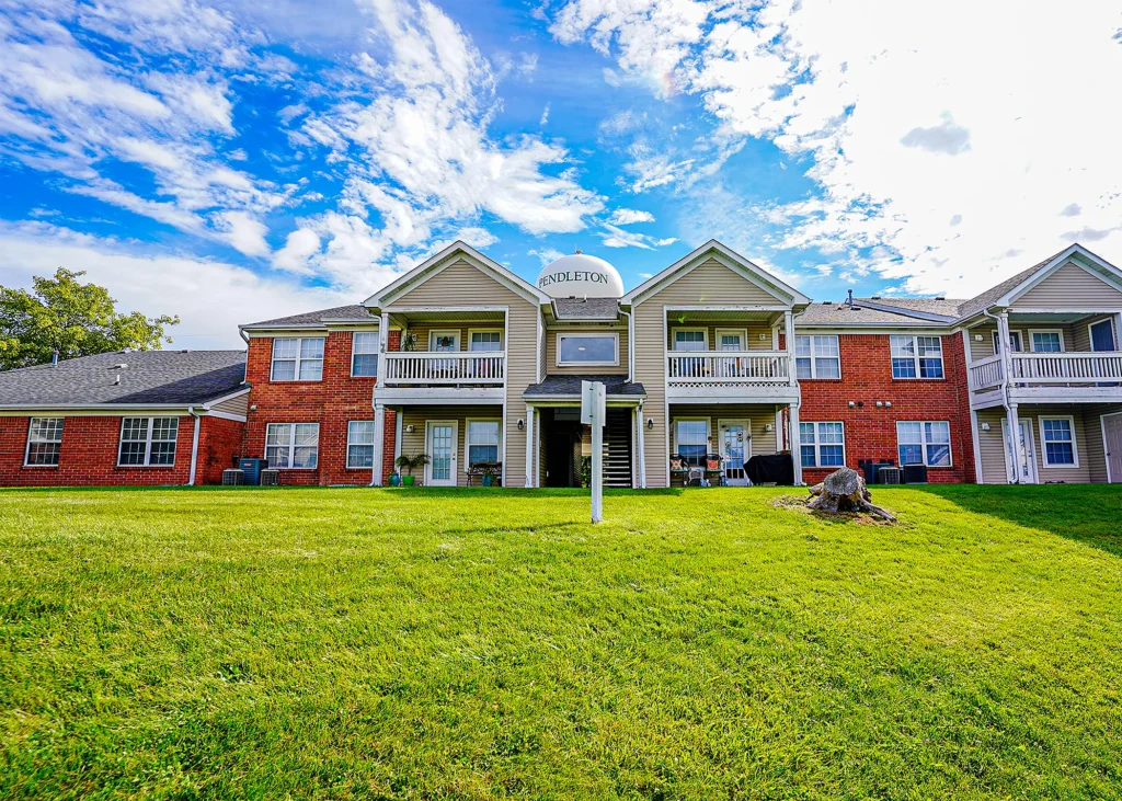A two-story apartment building with brick and siding exterior, balconies, and a sign reading Pendleton on a grassy lawn under a partly cloudy blue sky.