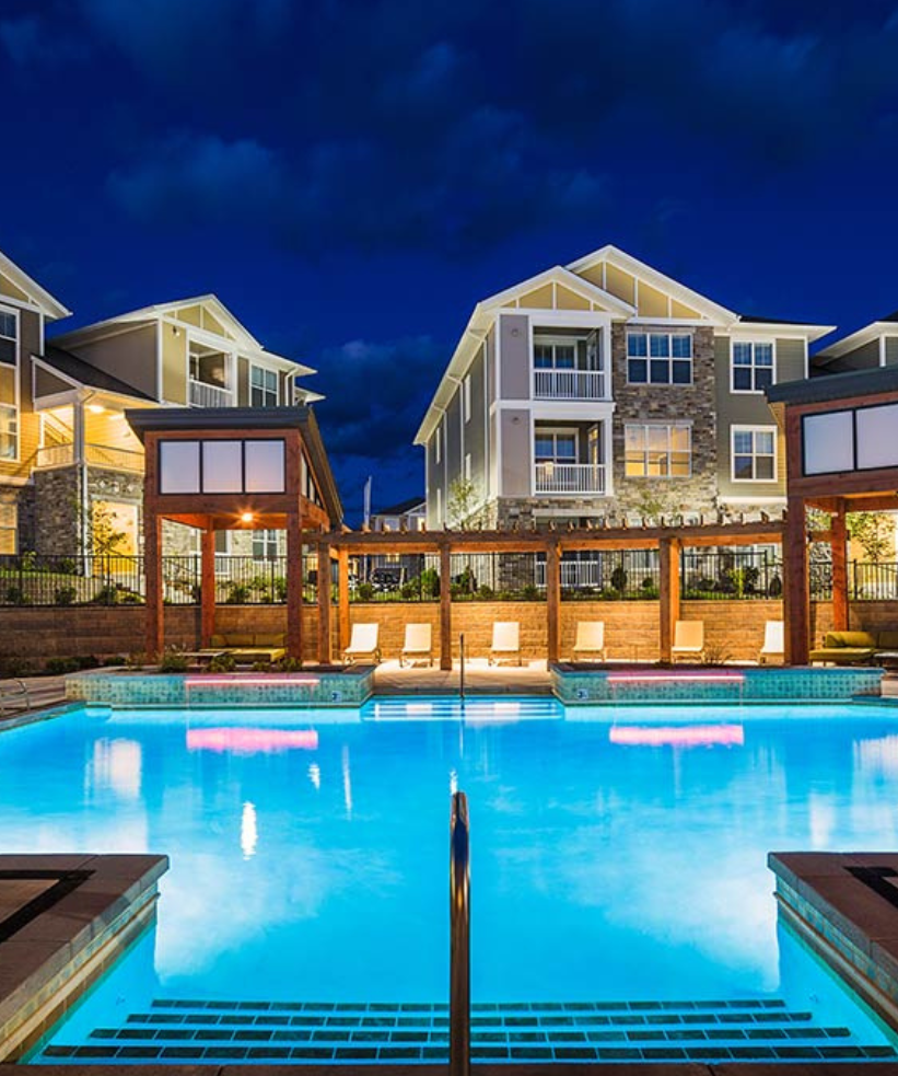 A brightly lit outdoor swimming pool at night, surrounded by lounge chairs and modern apartment buildings with large windows, under a dark, cloudy sky.
