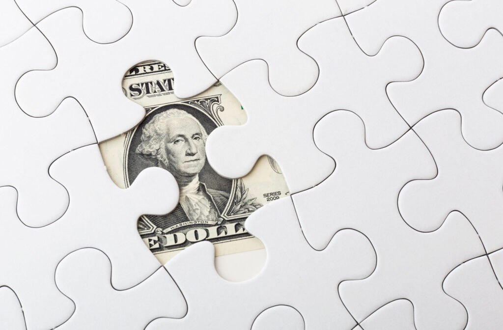 A close-up of a white jigsaw puzzle with one piece missing, revealing part of a U.S. one-dollar bill beneath the gap.