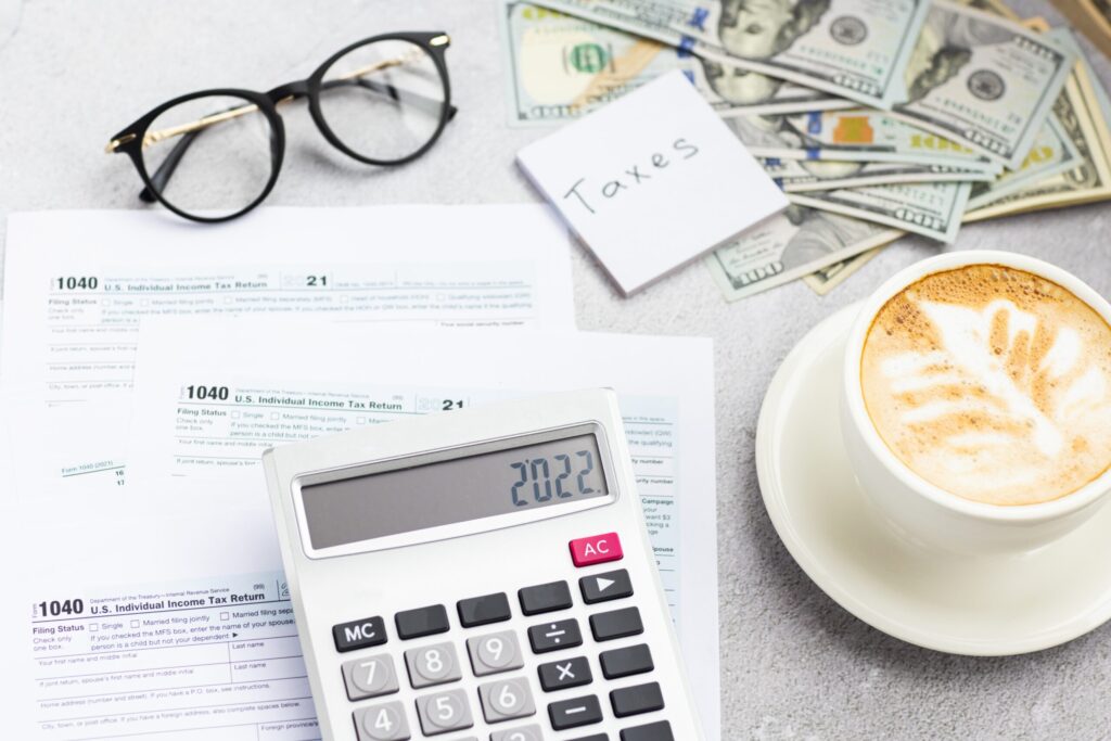 A calculator, eyeglasses, tax forms, a stack of cash, a note labeled Taxes, and a cup of cappuccino with heart latte art are arranged on a light surface.