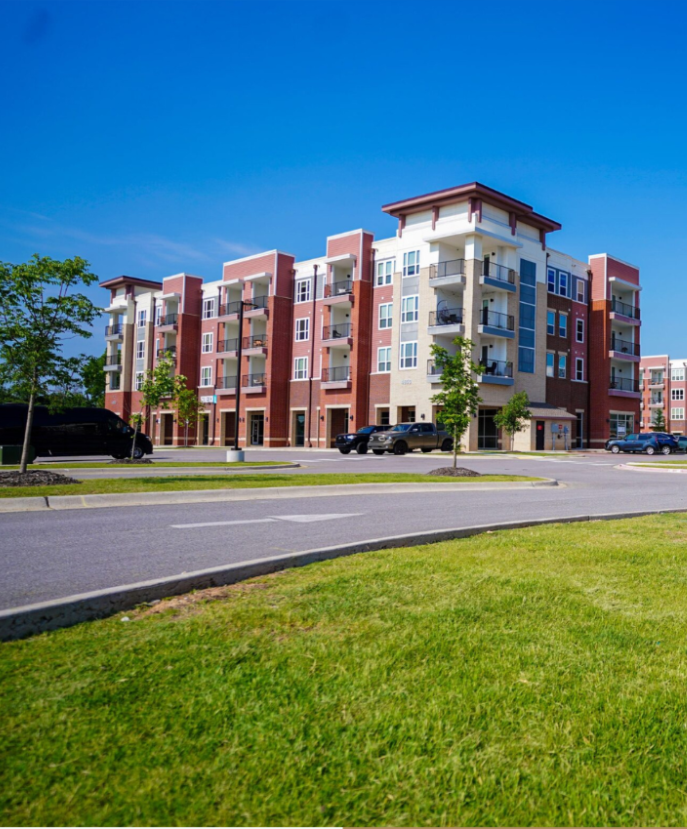 A modern four-story apartment building with red and beige exterior, balconies, and large windows. Several cars are parked in front, and theres a curved driveway and green lawn under a clear blue sky.