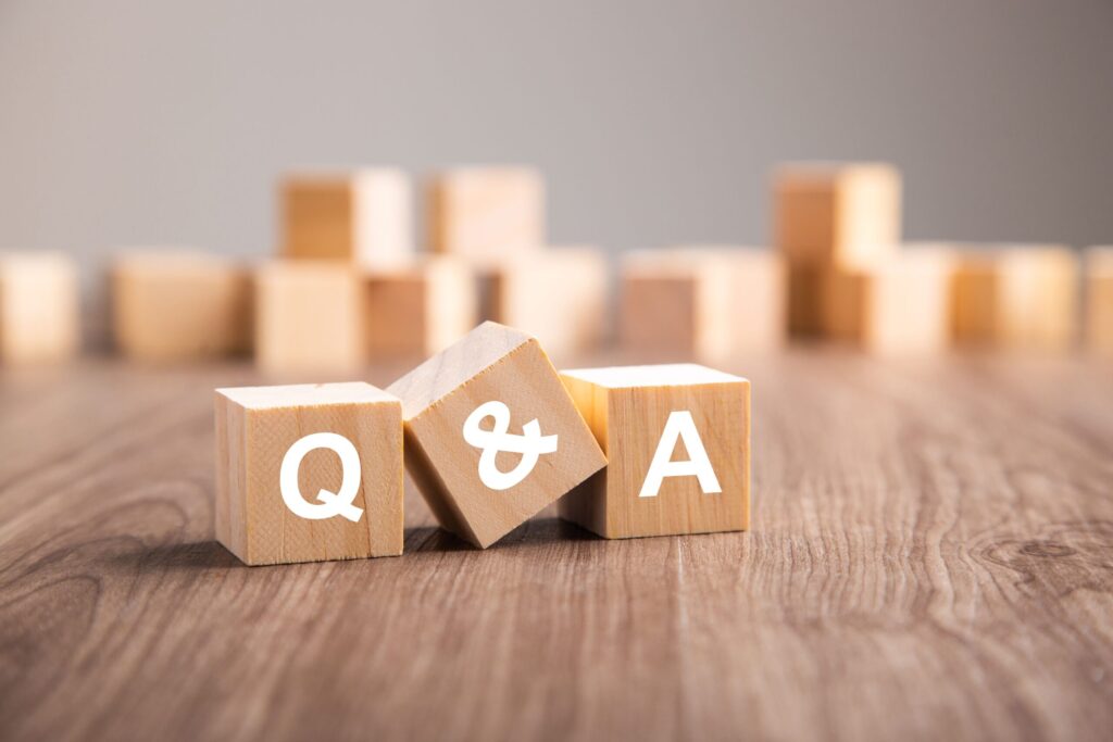 Three wooden blocks with the letters Q, &, and A printed on them are arranged on a wooden surface, representing Q&A. More blurred blocks are in the background.