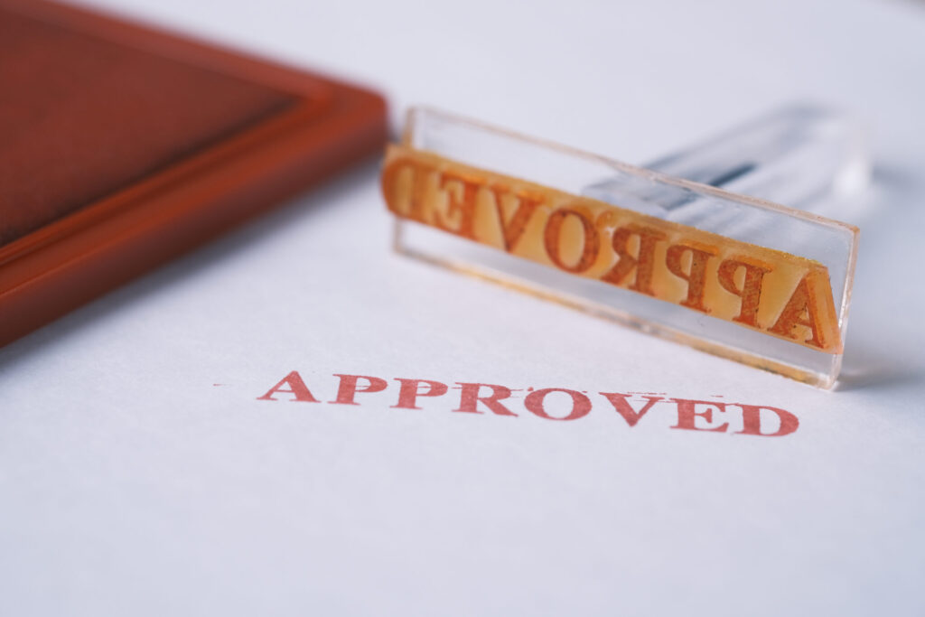 A close-up of a rubber stamp and ink pad. The stamp is upside down and reads APPROVED, matching the red APPROVED mark stamped on the white paper beneath it.