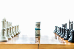 A roll of US dollar bills stands upright between white and black chess pieces set up for a game, symbolizing the connection between money and strategy or competition.