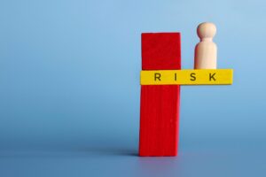 A wooden figure stands on the edge of a red block with a yellow plank labeled “RISK” against a blue background, symbolizing taking risks or facing danger.