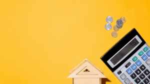 A small wooden house model, coins, and a calculator are arranged on a yellow background, suggesting a theme of home budgeting or financial planning.
