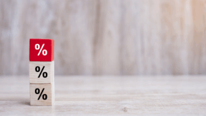 Three wooden blocks with percentage signs are stacked on top of each other on a light wooden surface. The top block is red, while the two below are natural wood color. The background is blurred and neutral.